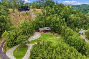 This aerial view showcases the property nestled in the lush, rolling hills of a forested mountain region. Multiple cabins are positioned among verdant trees, with scenic green spaces and access roads 