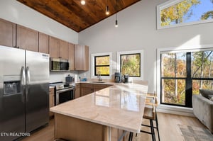 Step into your sunlit kitchen with soaring woodbeam ceilings and peaceful forest views framing every window. The bright island and modern appliances invite you to cook, gather, and soak in the natura