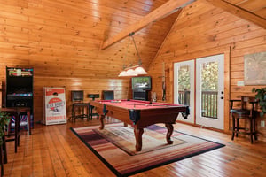 Your private game room awaits with a pool table and arcade entertainment beneath soaring timberbeam ceilings, opening onto your own balcony surrounded by trees.