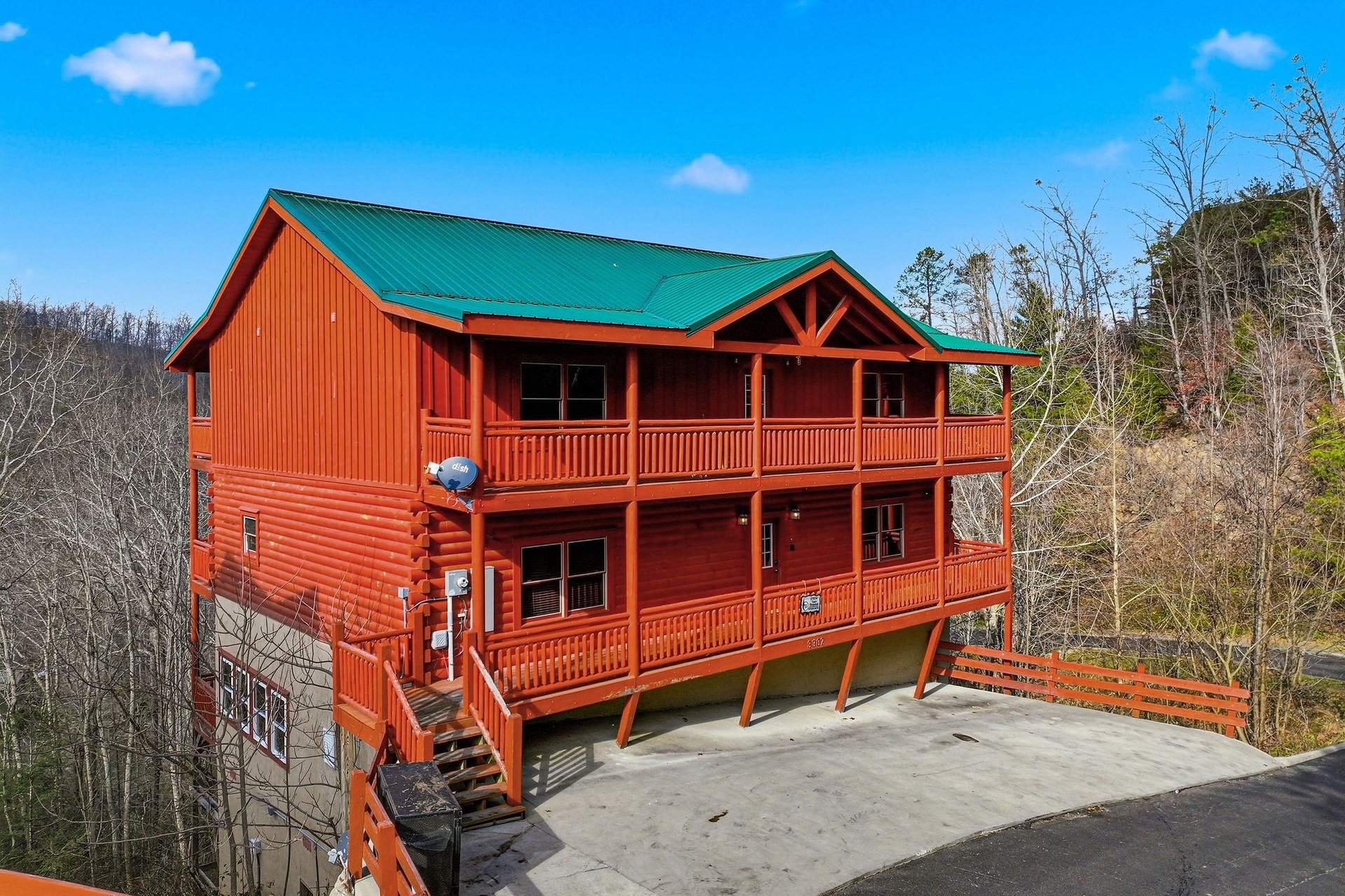 Stunning log cabin with green metal roof and wraparound decks nestled among rolling hills and forest landscape.