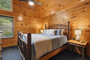 Step into your cozy cabin bedroom where rustic wood walls surround a handcrafted log bed layered with plush linens. Natural light filters through large windows, and a ceiling fan ensures your perfect 
