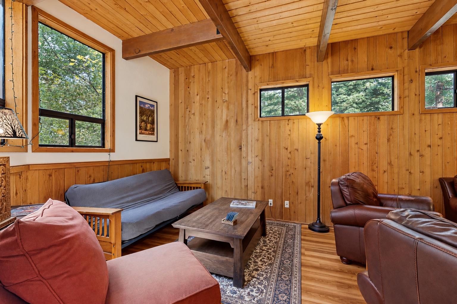 A relaxed second seating nook off the main living area, with knotty pine walls and forest views. The futon doubles as an extra sleeping spot for the group — just ask and well have sheets ready for it.