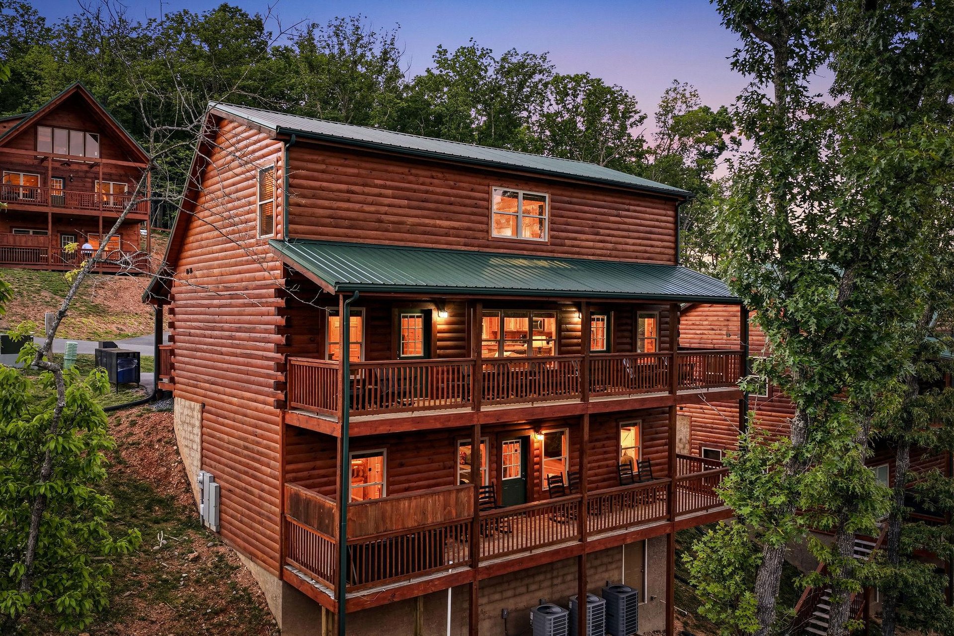 A welcoming mountain lodge featuring warm log construction, expansive decks on multiple levels, and softly lit interiors that glow as evening descends over the wooded hillside retreat.