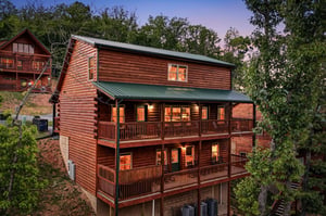A welcoming mountain lodge featuring warm log construction, expansive decks on multiple levels, and softly lit interiors that glow as evening descends over the wooded hillside retreat.