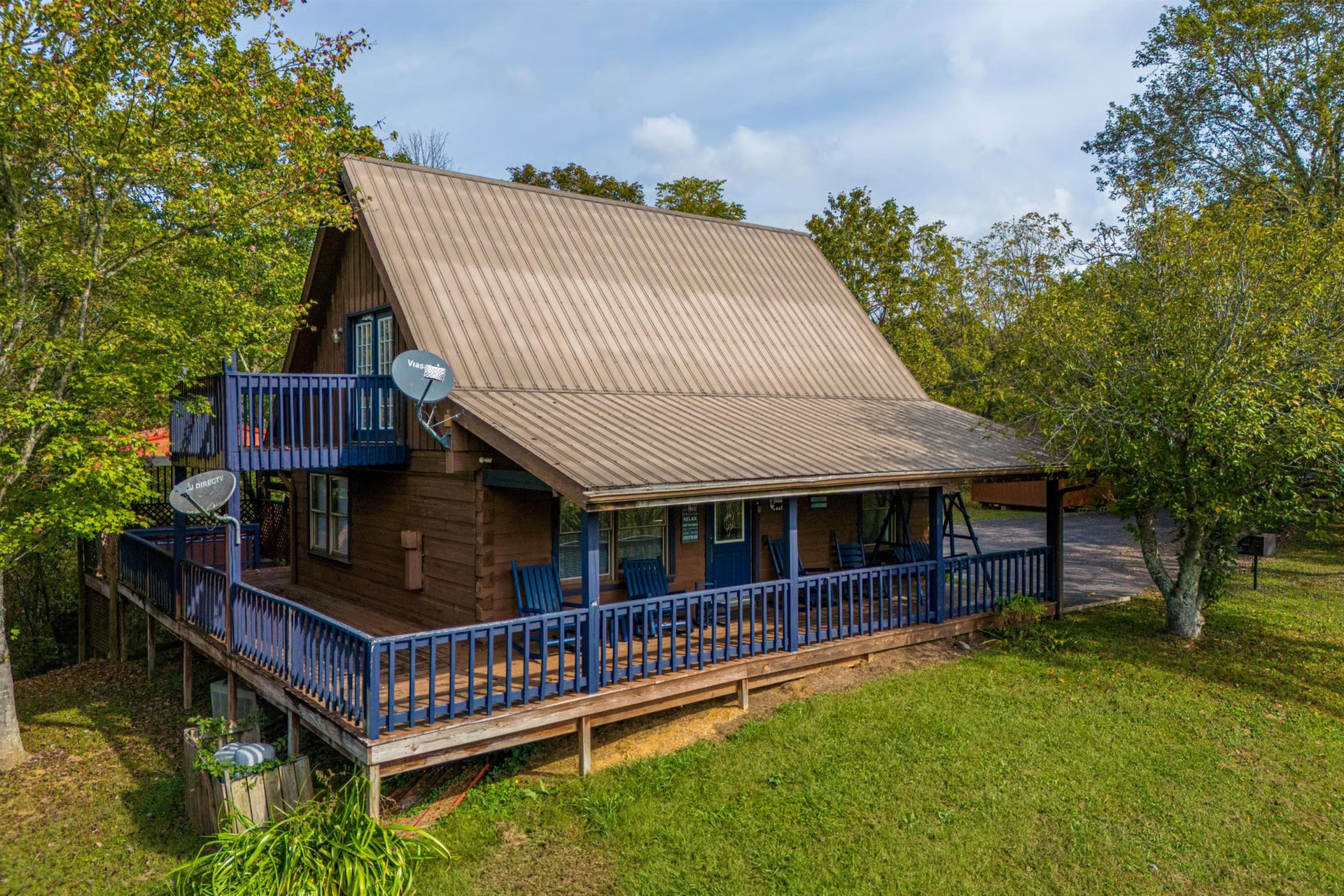 Charming Aframe cabin nestled among mature trees, featuring wraparound deck with blue railings and natural forest surroundings.