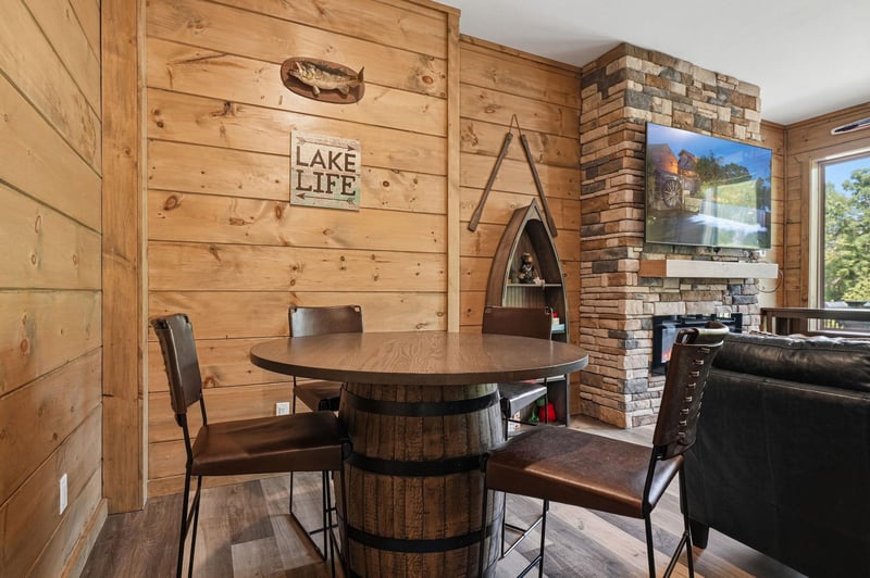 Gather around this rustic barrel dining table where warm wood walls and stone fireplace create your perfect lakeside retreat atmosphere.