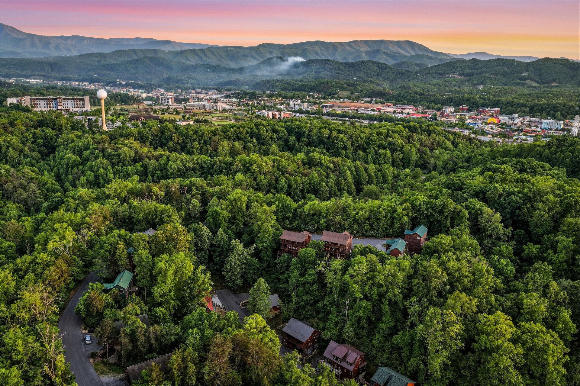 Spectacular aerial view of mountain resort cabins nestled among lush forests, with stunning sunset colors painting the sky over distant peaks.
