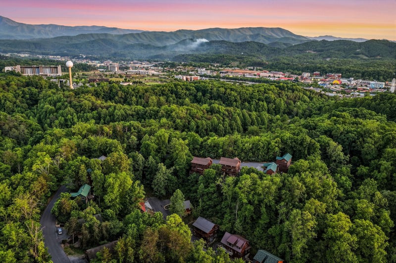 Spectacular aerial view of mountain resort cabins nestled among lush forests, with stunning sunset colors painting the sky over distant peaks.