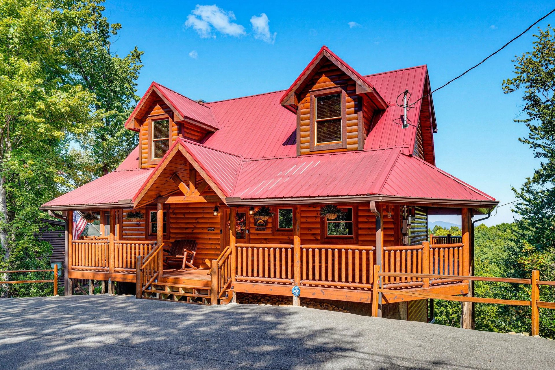A charming log cabin with a vibrant red roof, nestled among lush greenery and offering a welcoming wraparound porch beneath clear blue skies.