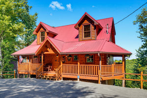 A charming log cabin with a vibrant red roof, nestled among lush greenery and offering a welcoming wraparound porch beneath clear blue skies.