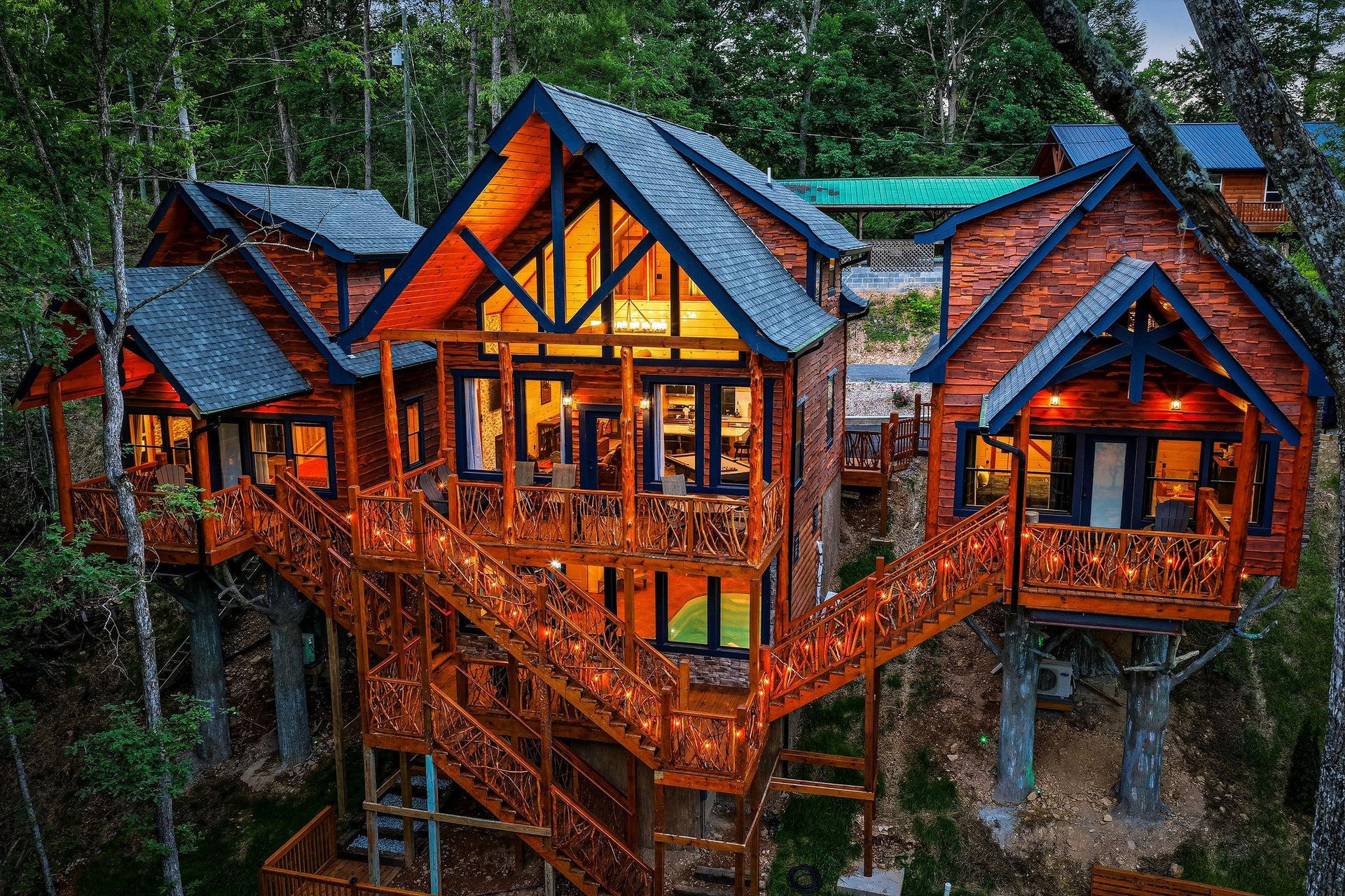 Stunning elevated property features rich wood siding, dramatic peaked windows, and wraparound decks nestled among towering trees at twilight. The illuminated interior showcases the inviting warmth of