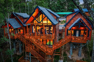 Stunning elevated property features rich wood siding, dramatic peaked windows, and wraparound decks nestled among towering trees at twilight. The illuminated interior showcases the inviting warmth of