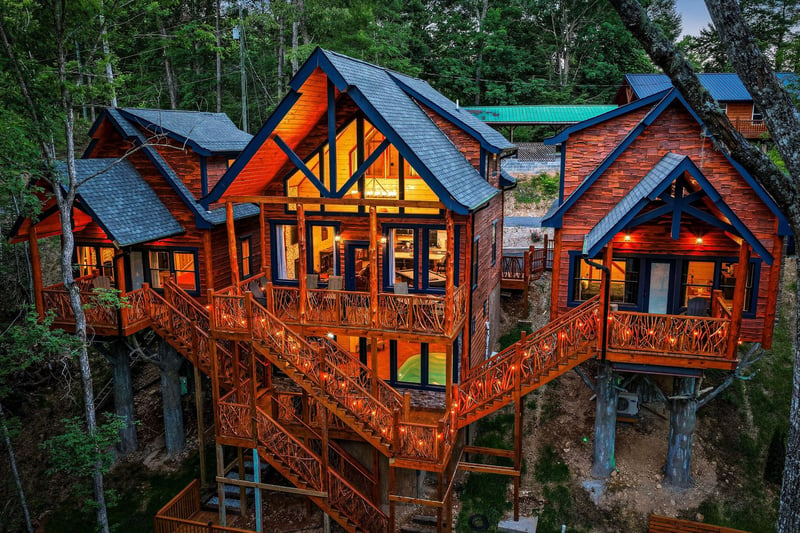 Stunning elevated property features rich wood siding, dramatic peaked windows, and wraparound decks nestled among towering trees at twilight. The illuminated interior showcases the inviting warmth of
