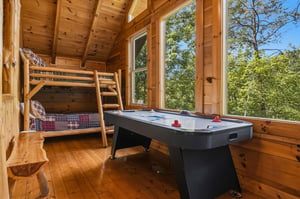 Your rustic retreat features a fun game room with a bunk bed and air hockey table, surrounded by warm wood and large windows framing the lush forest beyond.