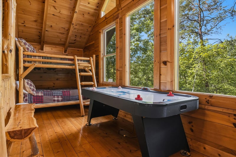 Your rustic retreat features a fun game room with a bunk bed and air hockey table, surrounded by warm wood and large windows framing the lush forest beyond.