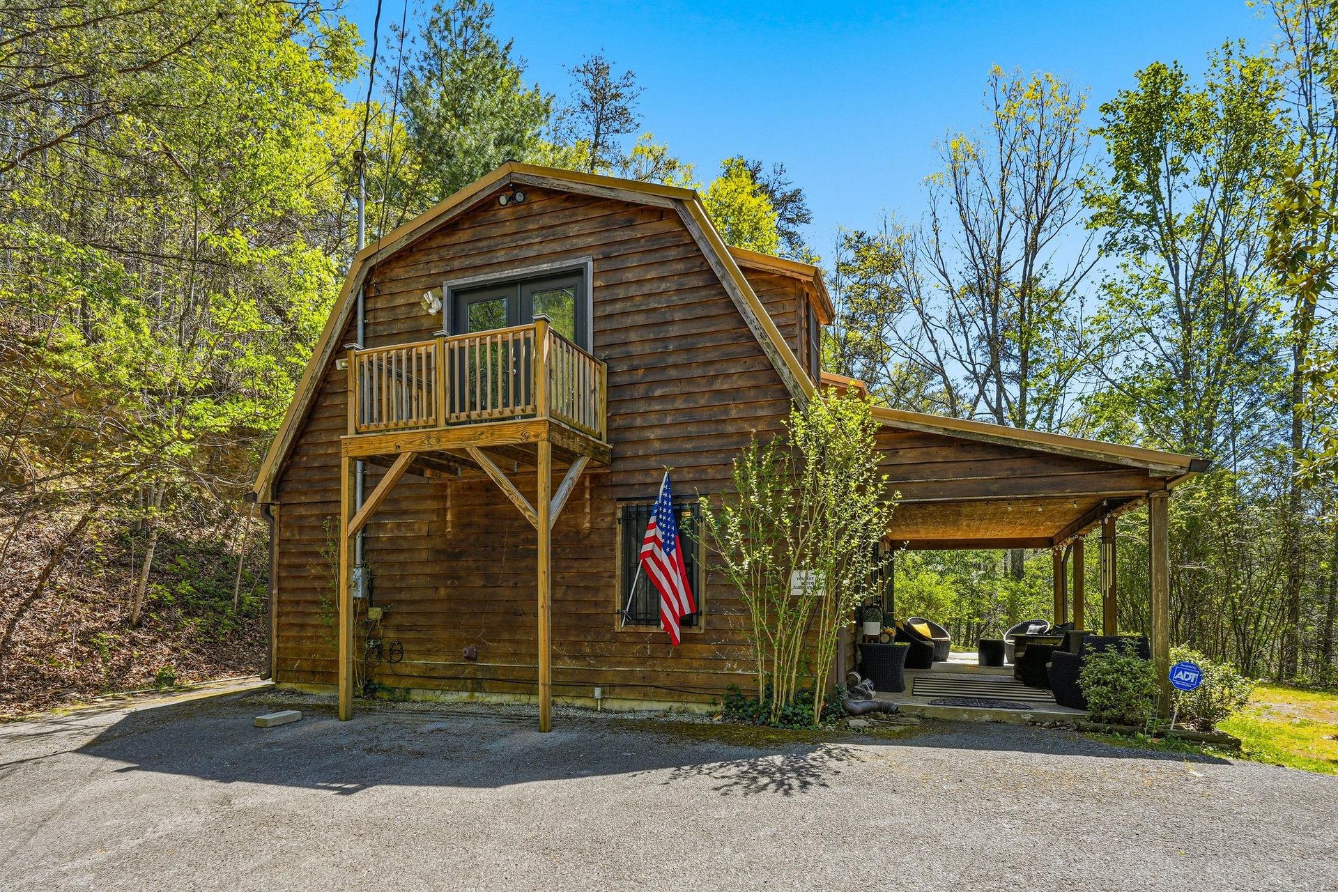 Charming wooden cabin surrounded by lush forest, featuring a private balcony and covered parking area.
