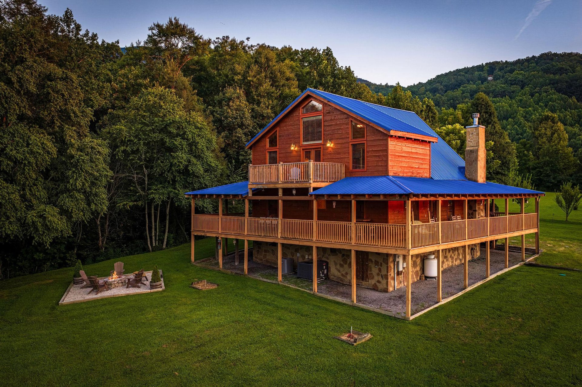 A striking mountain retreat features multiple wraparound decks and a welcoming fire pit area, set against rolling forested hills and open green space—perfect for peaceful outdoor evenings.