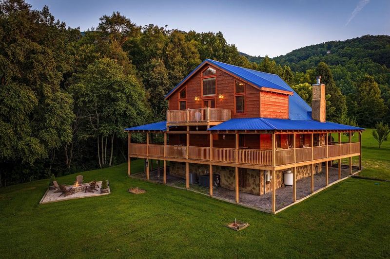 A striking mountain retreat features multiple wraparound decks and a welcoming fire pit area, set against rolling forested hills and open green space—perfect for peaceful outdoor evenings.