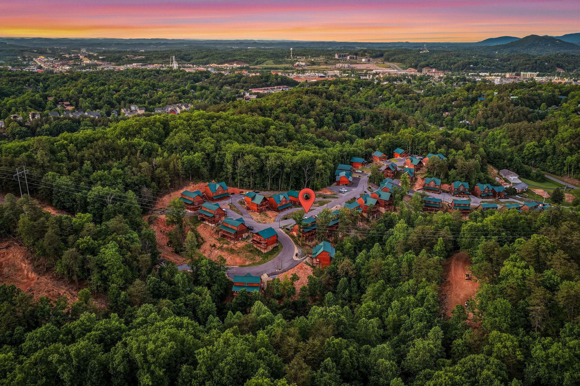 Stunning aerial view of the cabin resort community nestled in lush forests, with mountain vistas and vibrant sunset skies painting the horizon over the charming neighborhood.