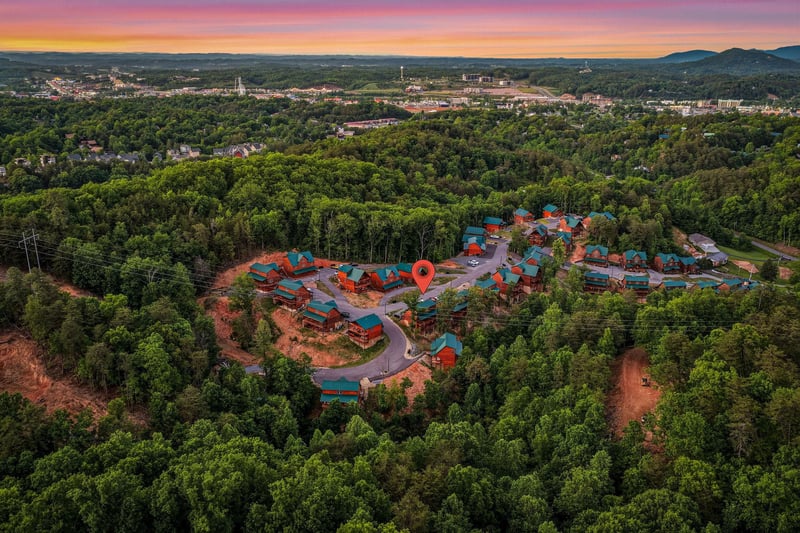 Stunning aerial view of the cabin resort community nestled in lush forests, with mountain vistas and vibrant sunset skies painting the horizon over the charming neighborhood.