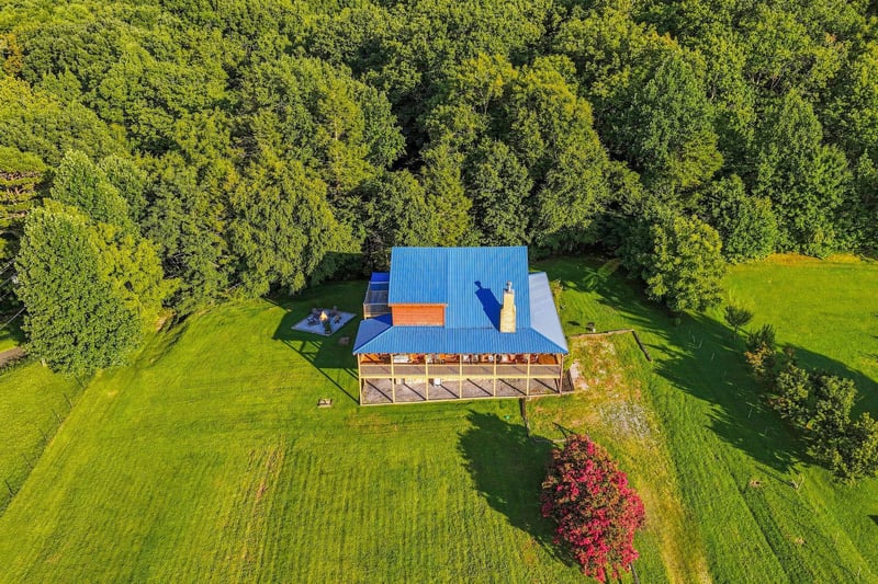 A charming property nestled on spacious grounds, surrounded by lush forest and vibrant seasonal foliage. The blue roof and wraparound deck stand out beautifully against the natural landscape.