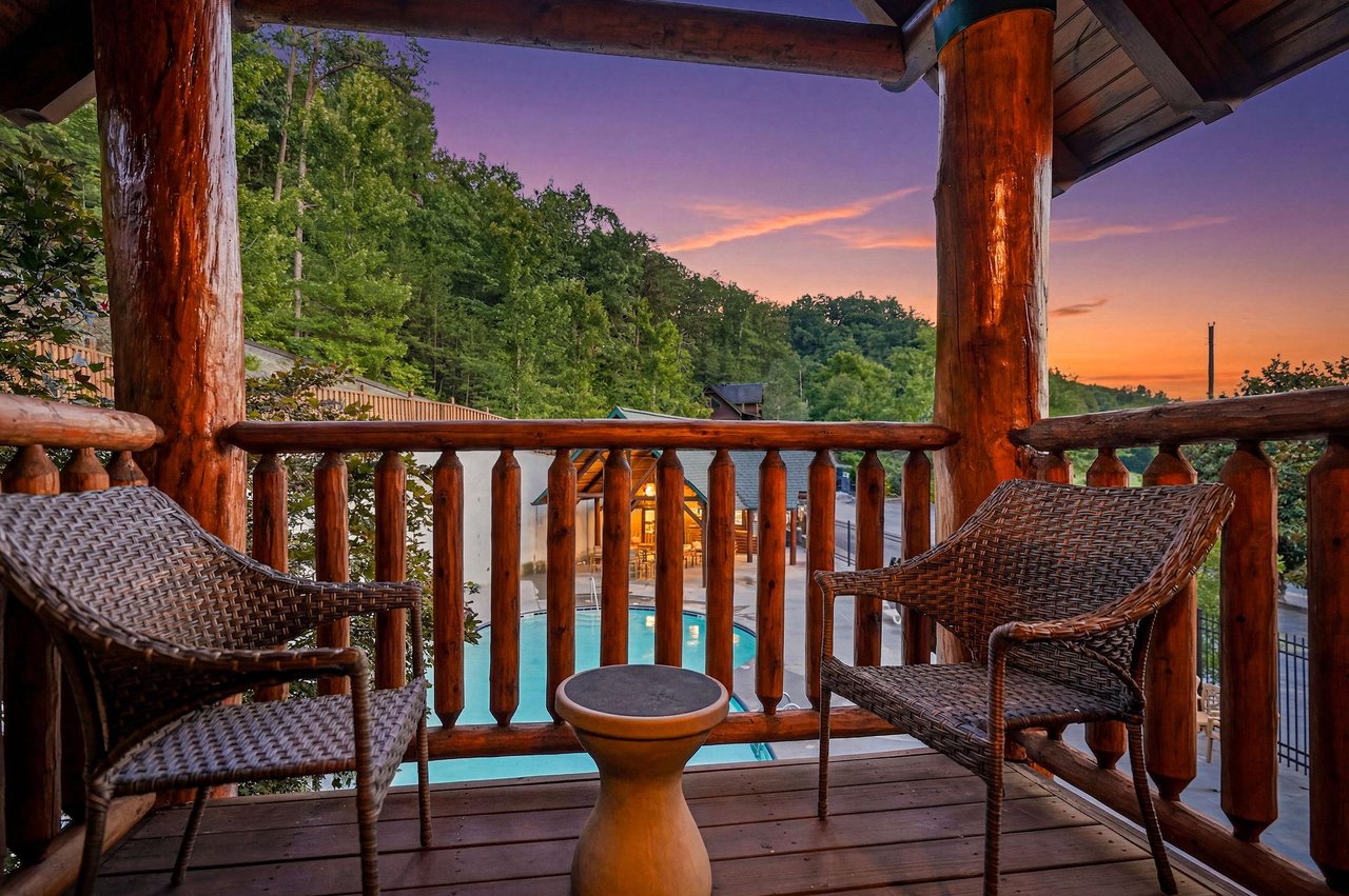 Sunset view from a private balcony overlooking the pool and natural landscape, framed by rustic log columns with comfortable wicker seating inviting peaceful mountain evenings.