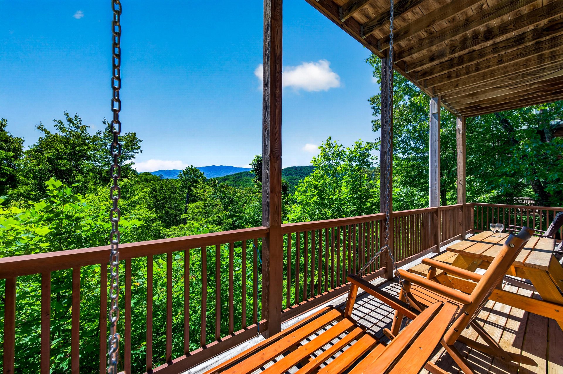 Expansive mountain panorama stretches before a covered terrace with swinging bench—lush treetops, rolling peaks, and clear skies frame this peaceful retreat.