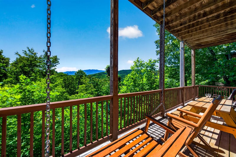 Expansive mountain panorama stretches before a covered terrace with swinging bench—lush treetops, rolling peaks, and clear skies frame this peaceful retreat.