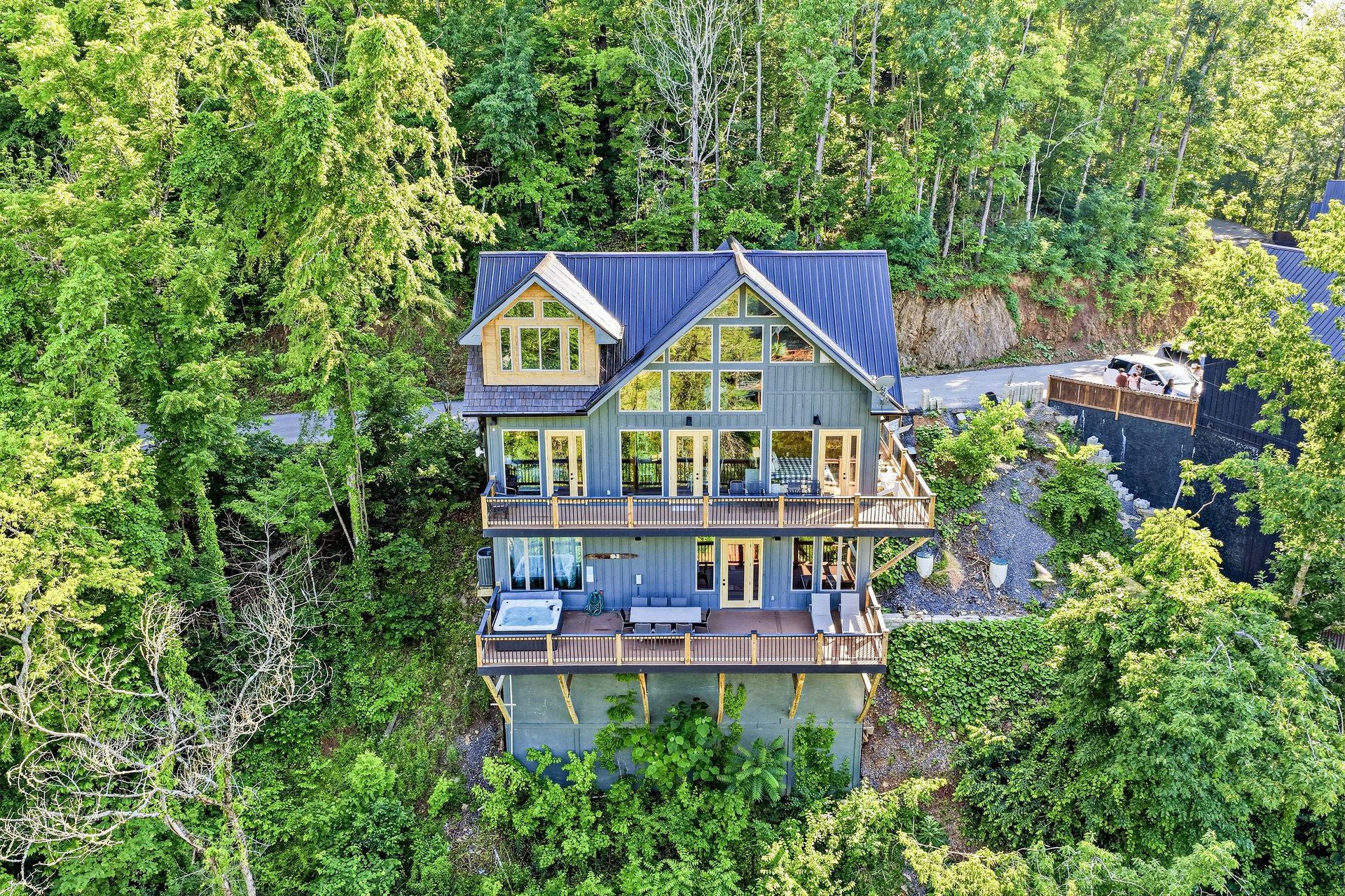 A striking modern mountain home nestled privately among lush forest, featuring expansive windows, multiple deck levels, and a blue metal roof that complements the surrounding natural landscape.