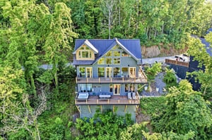 A striking modern mountain home nestled privately among lush forest, featuring expansive windows, multiple deck levels, and a blue metal roof that complements the surrounding natural landscape.