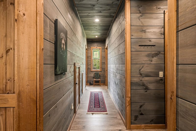 A warm hallway with rustic woodpaneled walls and ceiling leads to a peaceful nook, complete with a cozy reading chair and colorful runner beneath your feet.