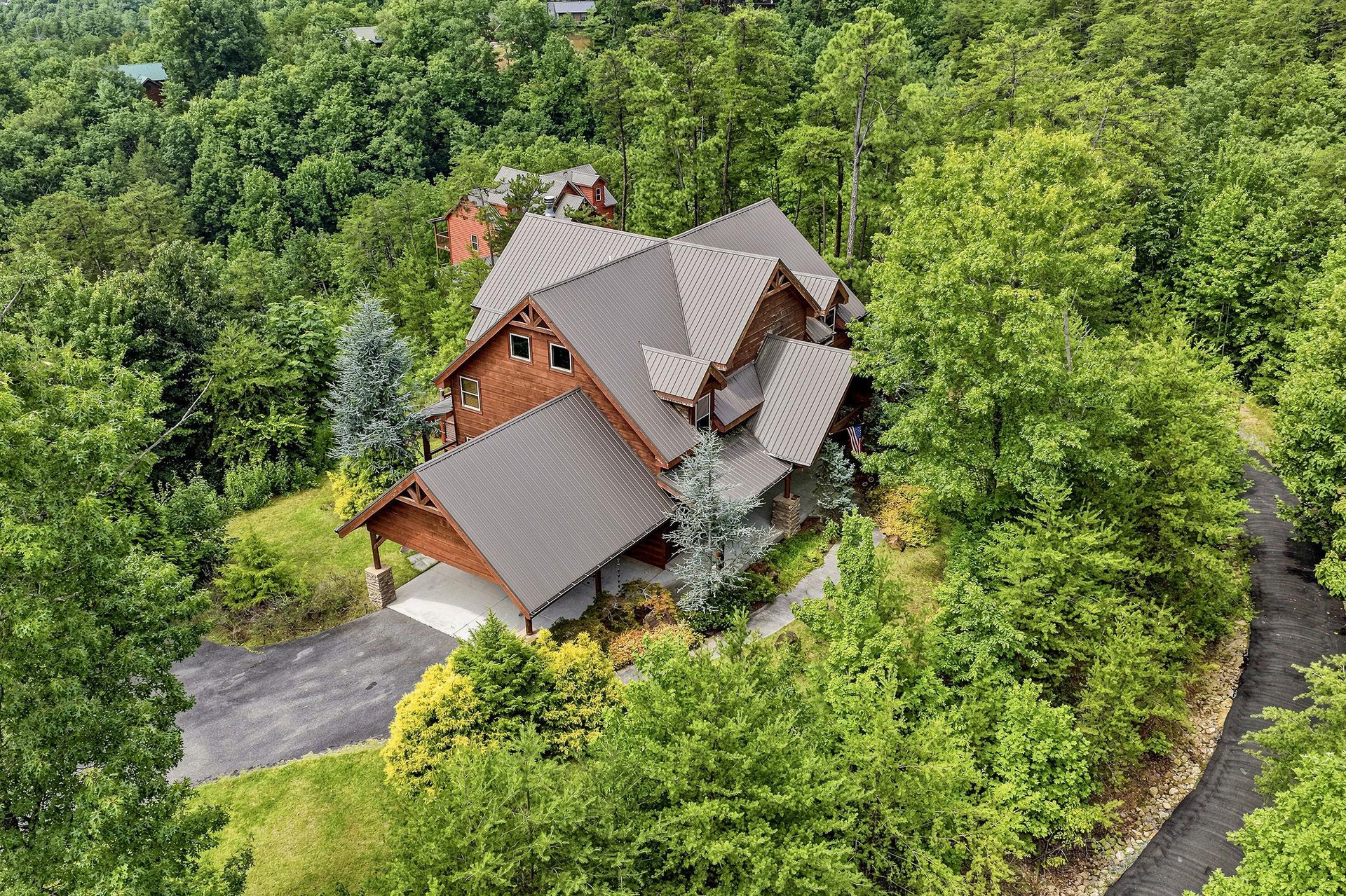Aerial view captures a spacious mountain lodge with distinctive multigabled roofing, surrounded by lush forested hillsides. The property features ample parking and landscaped grounds with mature tree