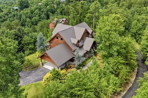 Aerial view captures a spacious mountain lodge with distinctive multigabled roofing, surrounded by lush forested hillsides. The property features ample parking and landscaped grounds with mature tree