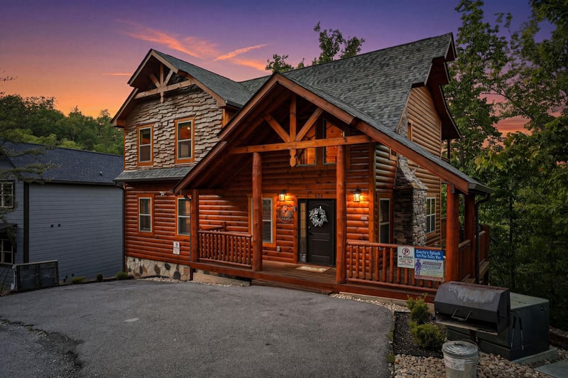 Stunning log cabin featuring rustic charm with stone accents, surrounded by lush trees under a breathtaking sunset sky.
