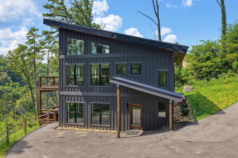 Modern architectural retreat nestled among lush greenery and tall trees. The property features sleek dark exterior siding, expansive windows, and multiple outdoor decks with scenic forest surroundings