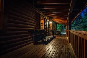 A charming log cabin porch invites evening relaxation with a classic swing and warm ambient lighting overlooking the surrounding wooded landscape.