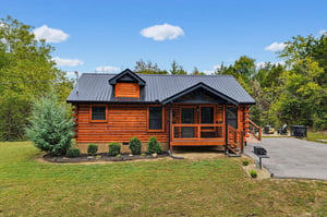 Surrounded by trees and landscaped grounds, this charming log cabin features a welcoming covered porch and sleek metal roof under blue skies.