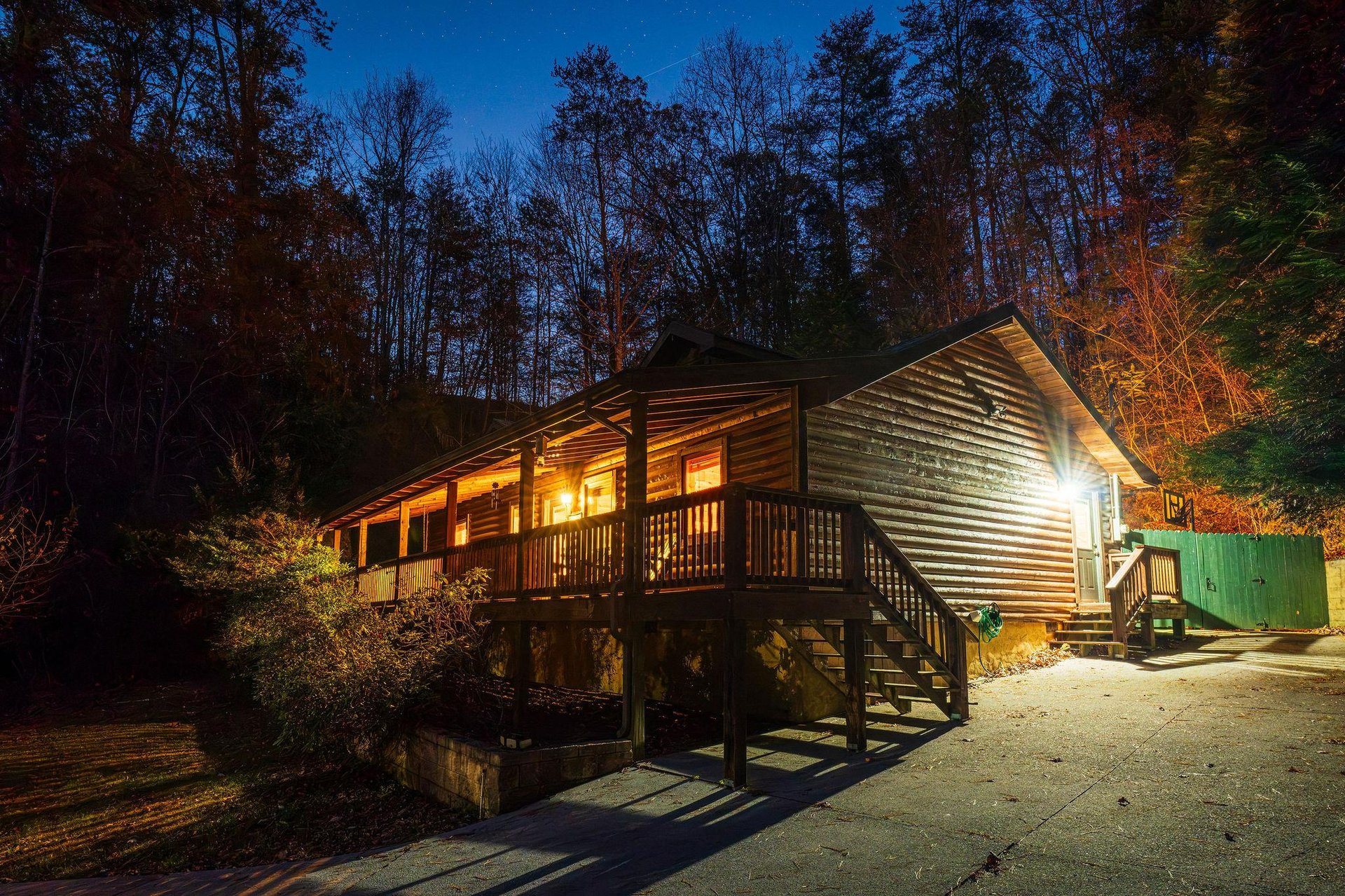 A cozy cabin nestled in the woods awaits, its warm lights glowing softly against the evening sky and towering trees that surround the property.