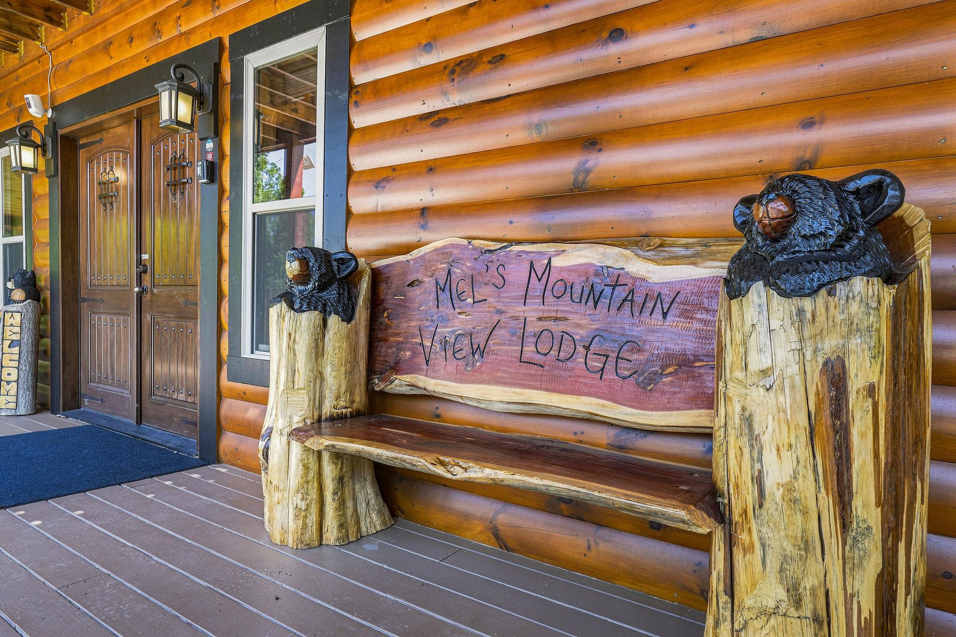 Mels Mountain View Lodge welcomes guests with a charming logcarved bench and handcarved bear statues at the entrance, setting a cozy mountain retreat tone.