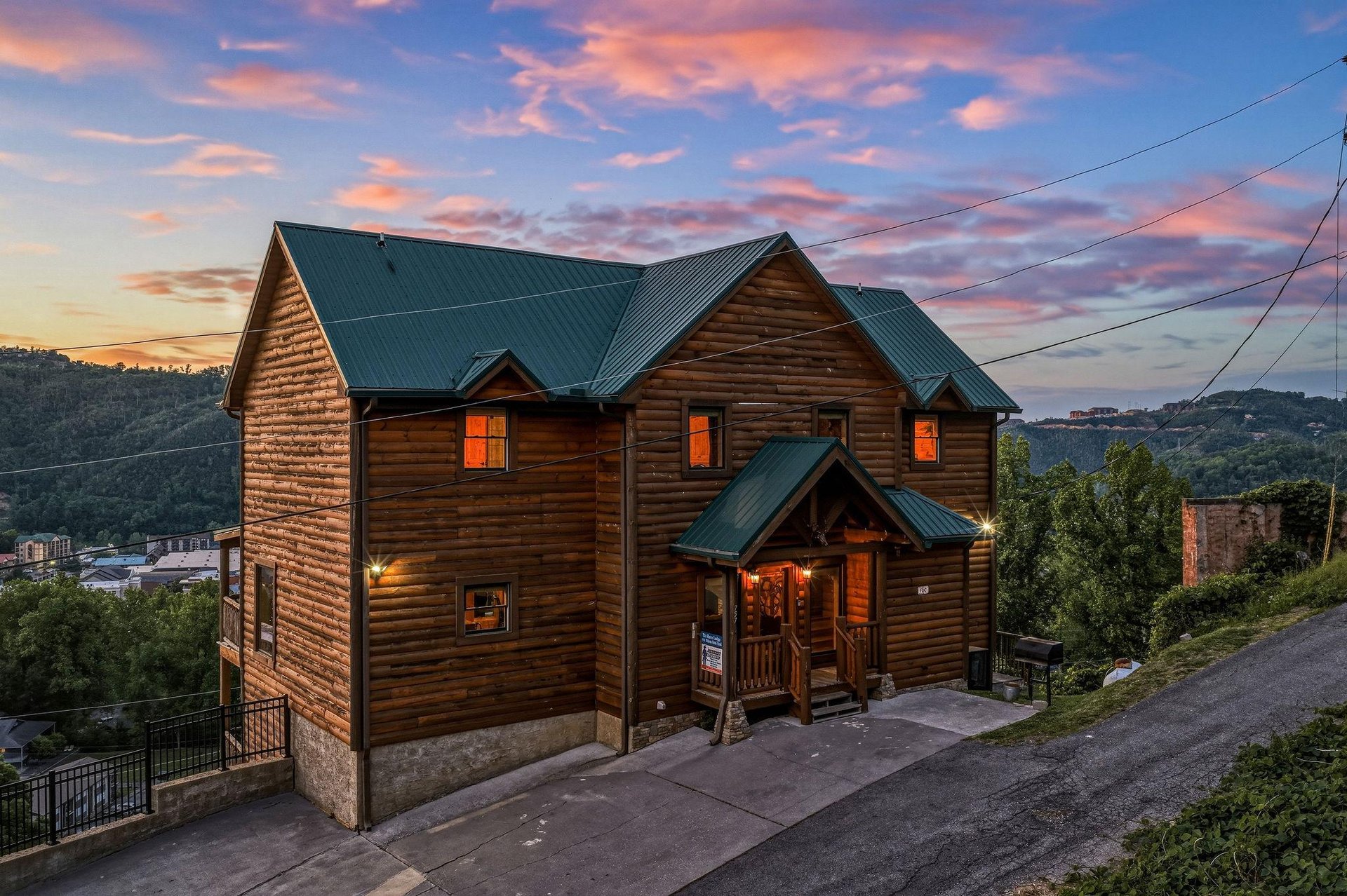 A charming log cabin nestled on a hillside, framed by rolling mountains and glowing warmly against a painted sunset sky in the surrounding area.