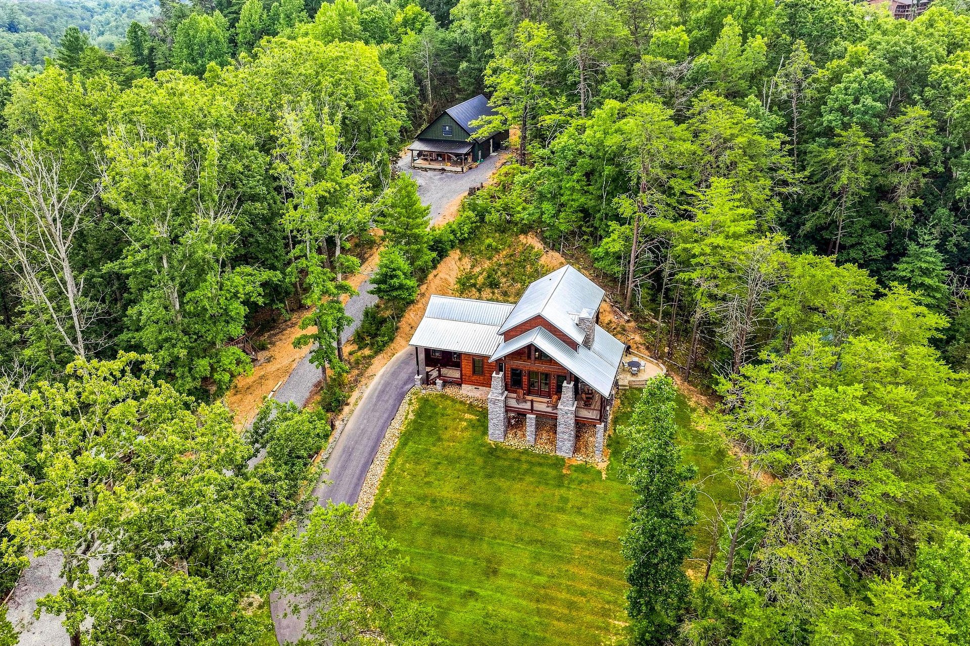 Aerial view of two mountain cabin rentals nestled in a lush forest setting, with paved driveways and private lawns surrounded by dense green woodland.
