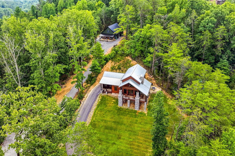 Aerial view of two mountain cabin rentals nestled in a lush forest setting, with paved driveways and private lawns surrounded by dense green woodland.