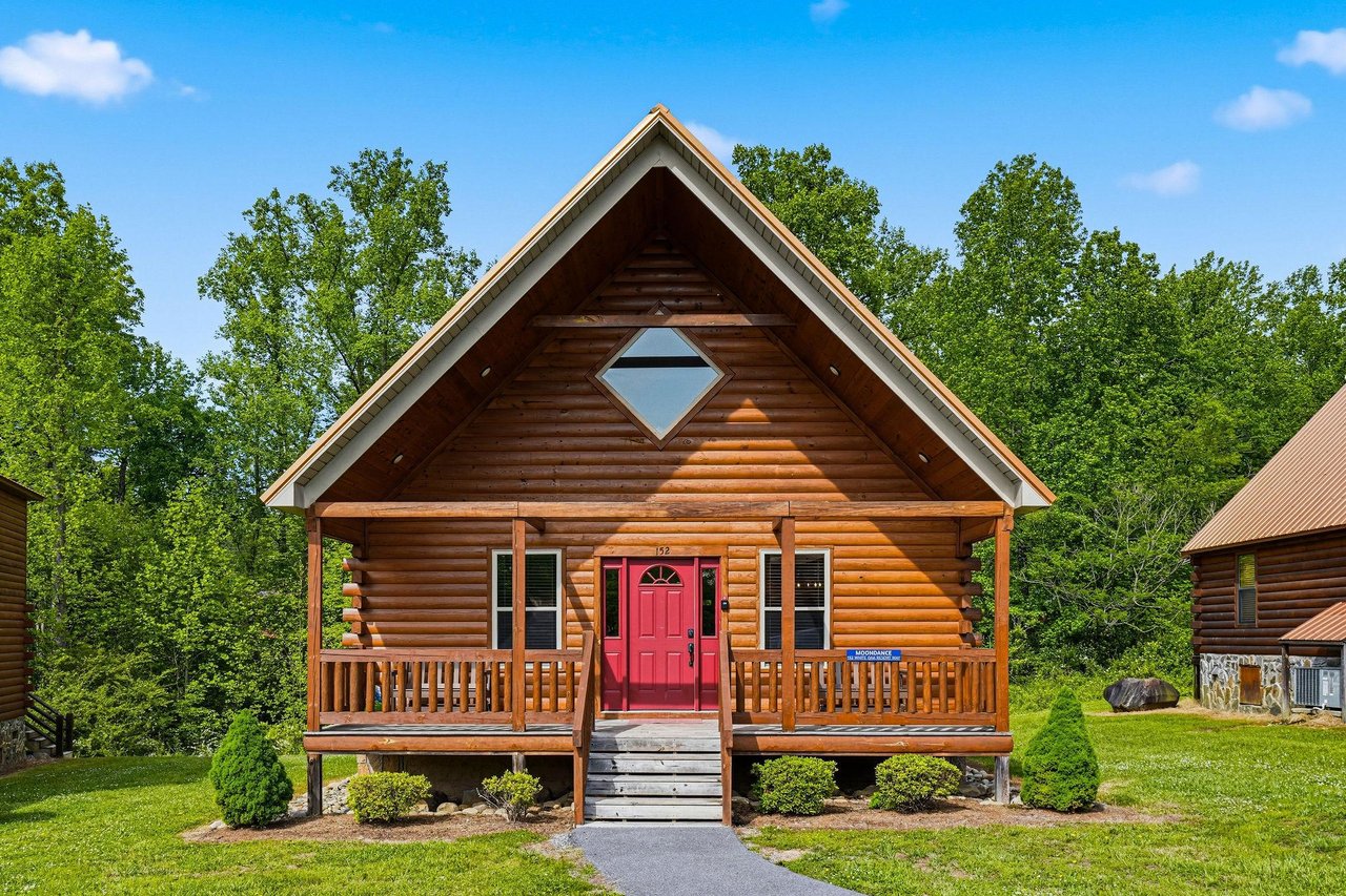 A charming log cabin with a welcoming red door and covered porch, nestled among lush green trees in a peaceful natural setting.