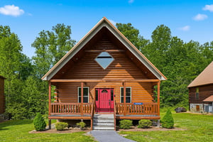 A charming log cabin with a welcoming red door and covered porch, nestled among lush green trees in a peaceful natural setting.