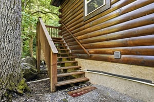 Rustic log cabin exterior featuring warm wood construction and an outdoor stairway, surrounded by mature forest trees creating a peaceful natural setting.