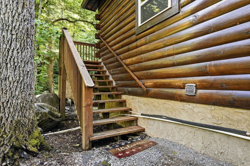 Rustic log cabin exterior featuring warm wood construction and an outdoor stairway, surrounded by mature forest trees creating a peaceful natural setting.