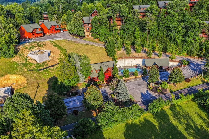 Aerial view of a wooded vacation rental community featuring rustic cabinstyle buildings, an outdoor swimming pool, mature landscaping, and winding paved roads through lush forest surroundings.