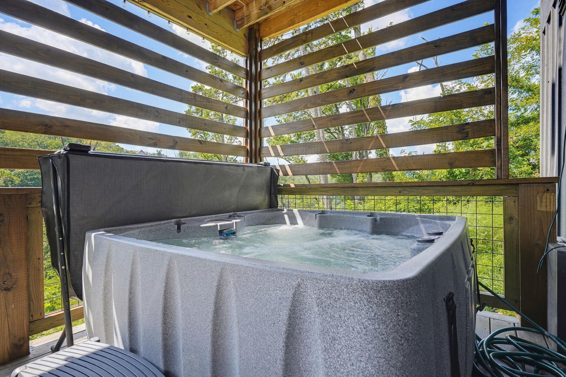 Soak in the warm, bubbling hot tub beneath a wooden pergola as sunlight filters through the slats and fresh greenery frames the horizon.