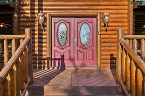 This stunning log cabin entrance features custom double doors with decorative glass panels, flanked by rustic log railings and welcoming lanternstyle sconces that frame your mountain retreat arrival.