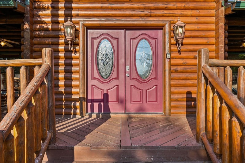 This stunning log cabin entrance features custom double doors with decorative glass panels, flanked by rustic log railings and welcoming lanternstyle sconces that frame your mountain retreat arrival.